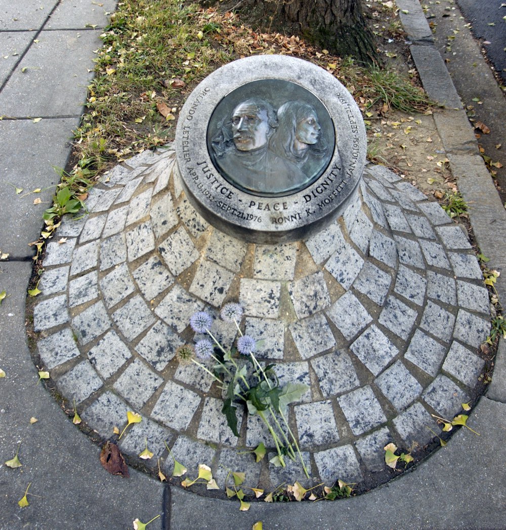 The Letelier-Moffitt memorial at Sheridan Circle, Washington D.C. — a low granite block engraved with the names 'Orlando Letelier 1932-1976' and 'Ronni Karpen Moffitt 1951-1976,' set into a paved pedestrian area at the edge of the traffic circle.