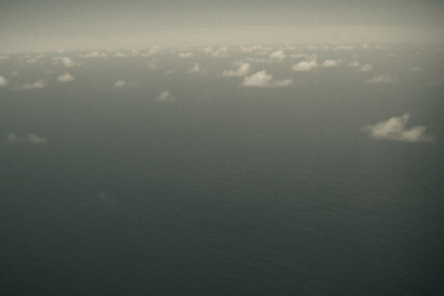 An empty Pacific Ocean horizon at midday seen from high altitude — calm dark-blue sea below, a thin scattered layer of mid-altitude cumulus broken by clear sky, no aircraft and no object in the frame.