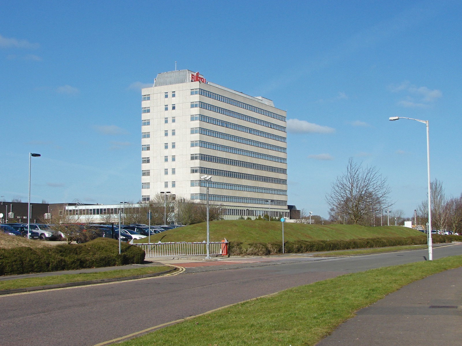 The Fujitsu building in Bracknell, Berkshire — a large low-rise modern office block of grey cladding and reflective glass set behind a car park and landscaped verges under an overcast sky.