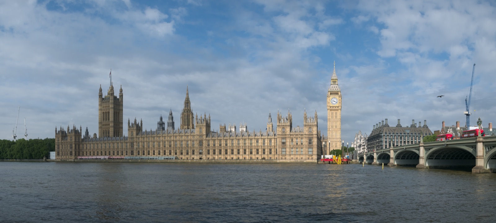 The Palace of Westminster and the Elizabeth Tower seen across the River Thames — the long Gothic Revival riverfront of the Houses of Parliament in honey-coloured stone, with the clock tower rising at the near end under a bright sky.