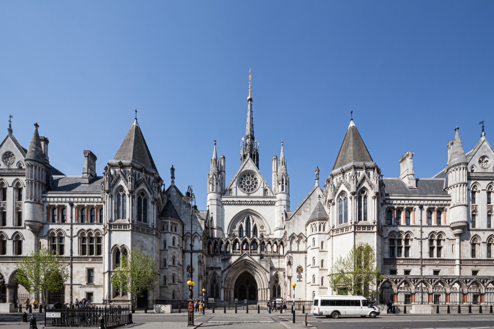 The Royal Courts of Justice on the Strand in London — a long Victorian Gothic Revival stone facade with a central spire, pointed arches, and twin towers, photographed from across the street under a clear blue sky.