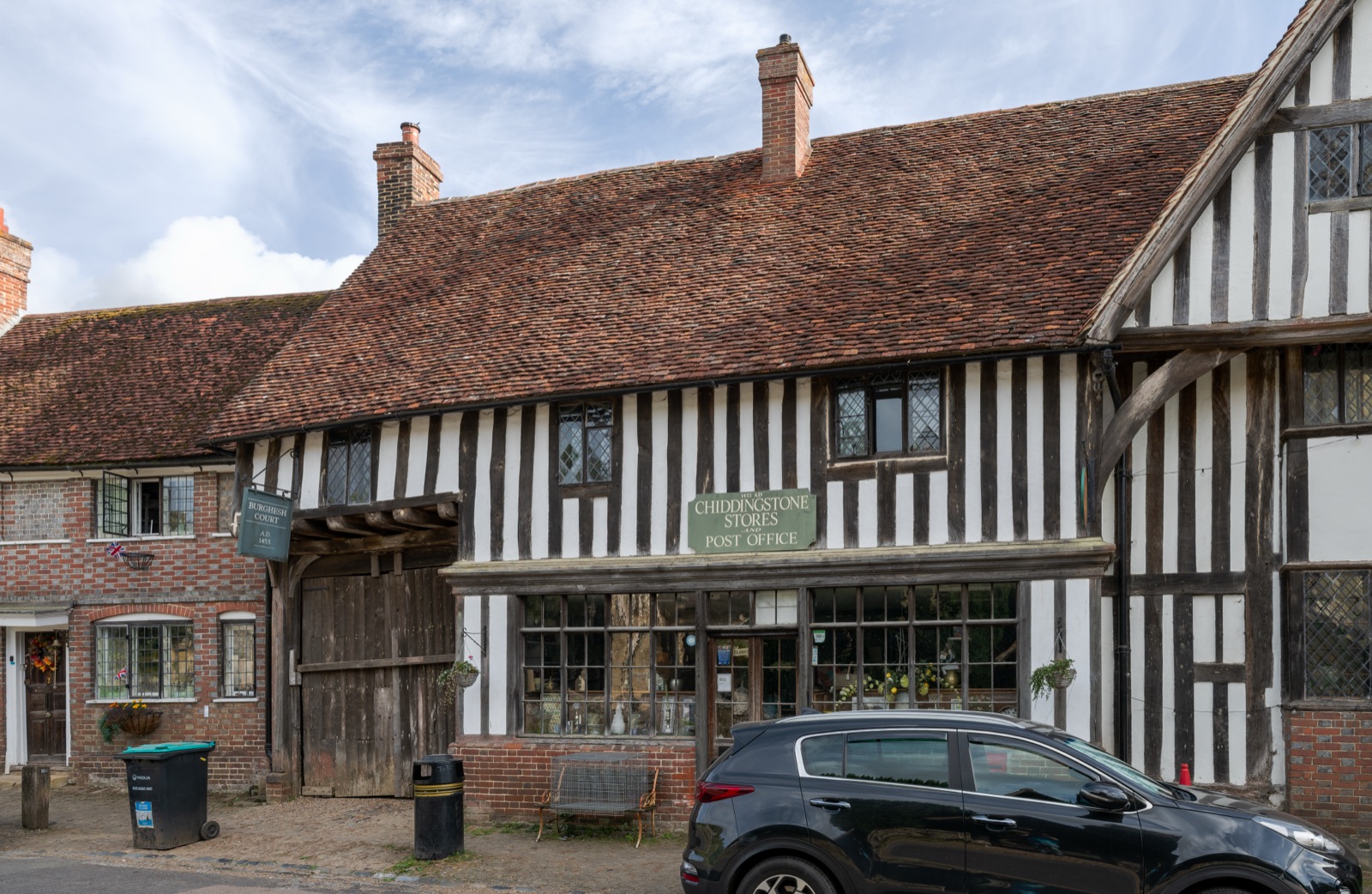 A timber-framed Tudor village shop and post office in Chiddingstone, Kent — a long black-and-white half-timbered building with a tiled roof, leaded shop windows, and a green sign reading 'Chiddingstone Stores Post Office', with a parked car in front.