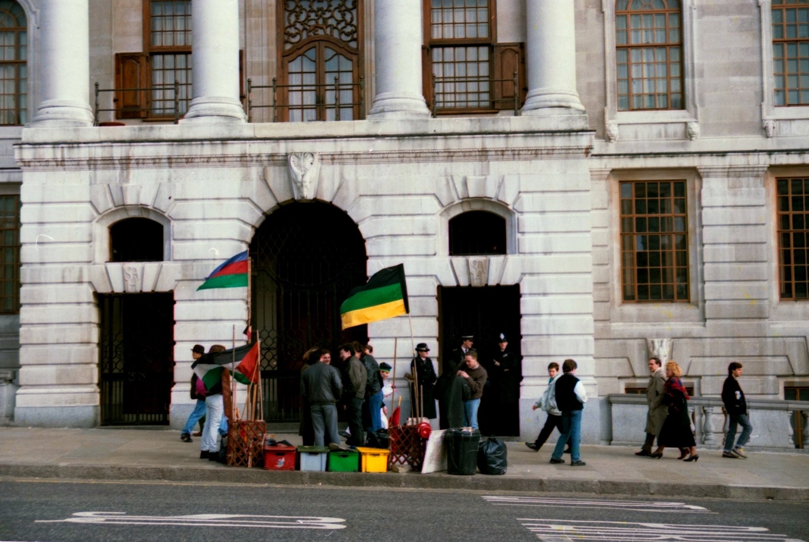 An anti-apartheid demonstration outside South Africa House in London in 1989: a small group of protesters with the green, black, and gold flags of the African National Congress gathered on the pavement in front of the grand stone building, watched by police.