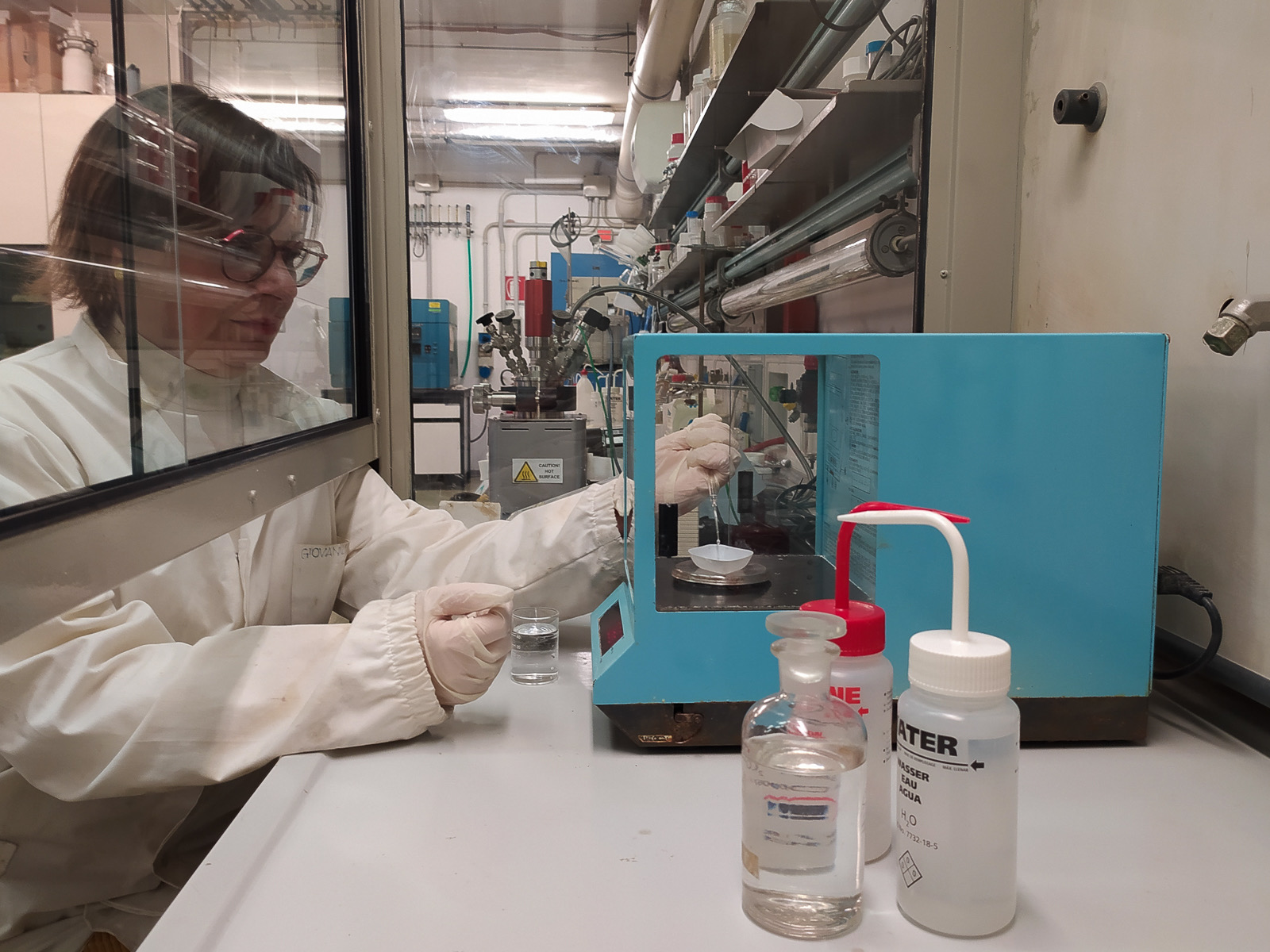 A modern chemistry laboratory fume hood with glassware and apparatus set up inside it, sashes raised, in a clean tiled lab.
