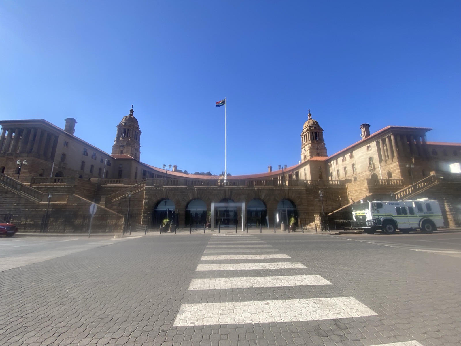 The Union Buildings in Pretoria, a long sandstone government complex with twin domed towers and curved colonnades, set above terraced gardens under a blue sky.