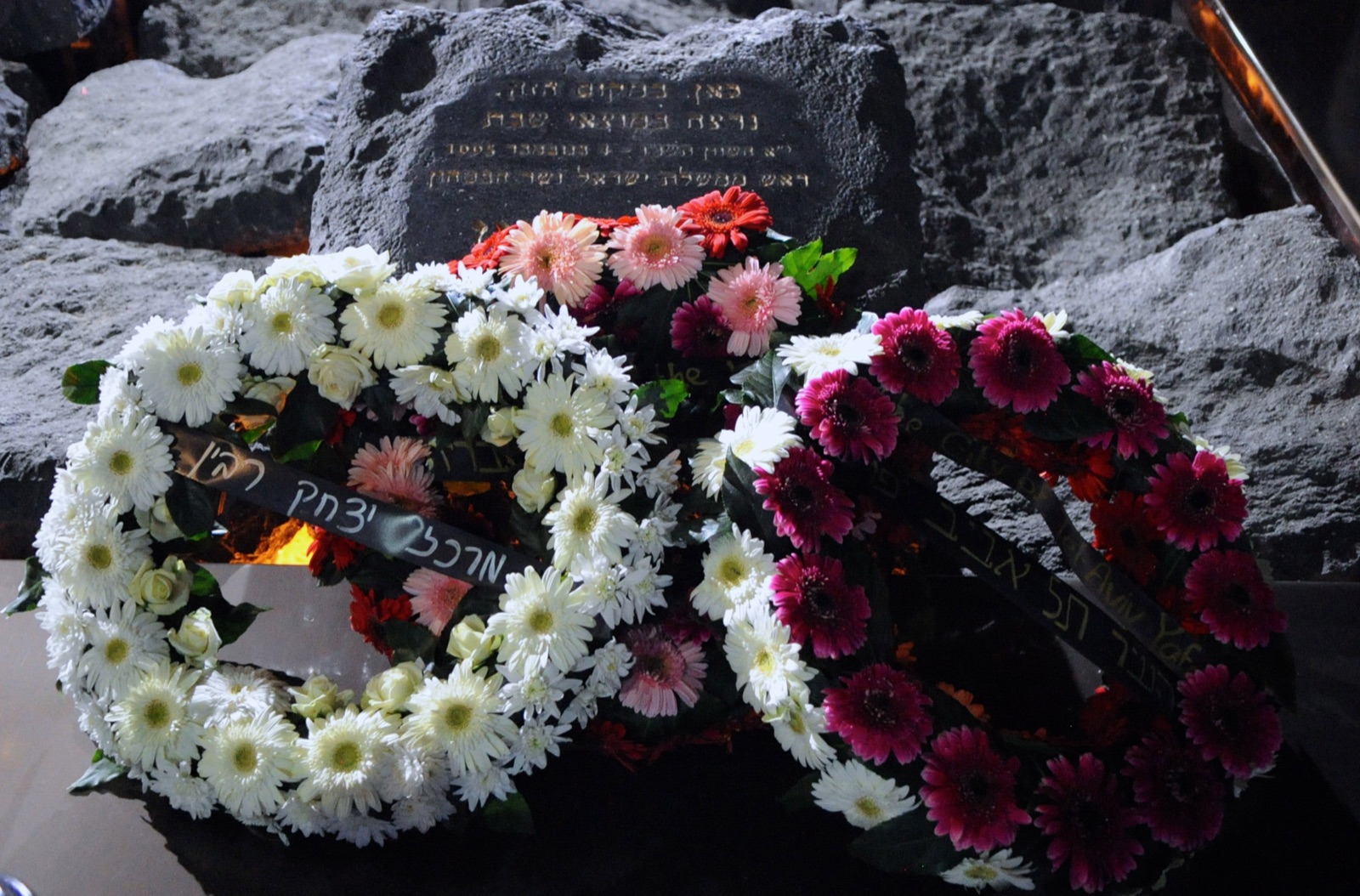 The Yitzhak Rabin memorial at the assassination site — rough black basalt boulders set into the ground around a dark inscribed memorial plaque in Hebrew, with several wreaths of white and deep-pink flowers laid against the stones.