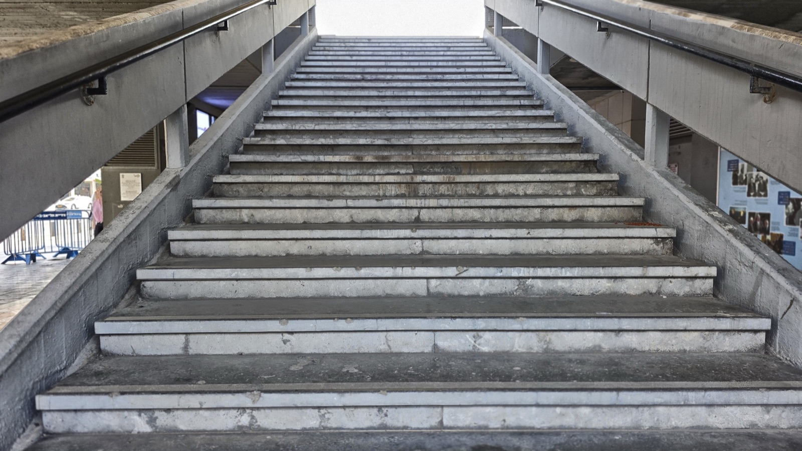 An empty exterior concrete staircase running up beneath the raised slab of Tel Aviv City Hall — broad bare steps between plain concrete walls and handrails, leading up toward bright daylight at the top, with part of the open plaza visible to the lower left.