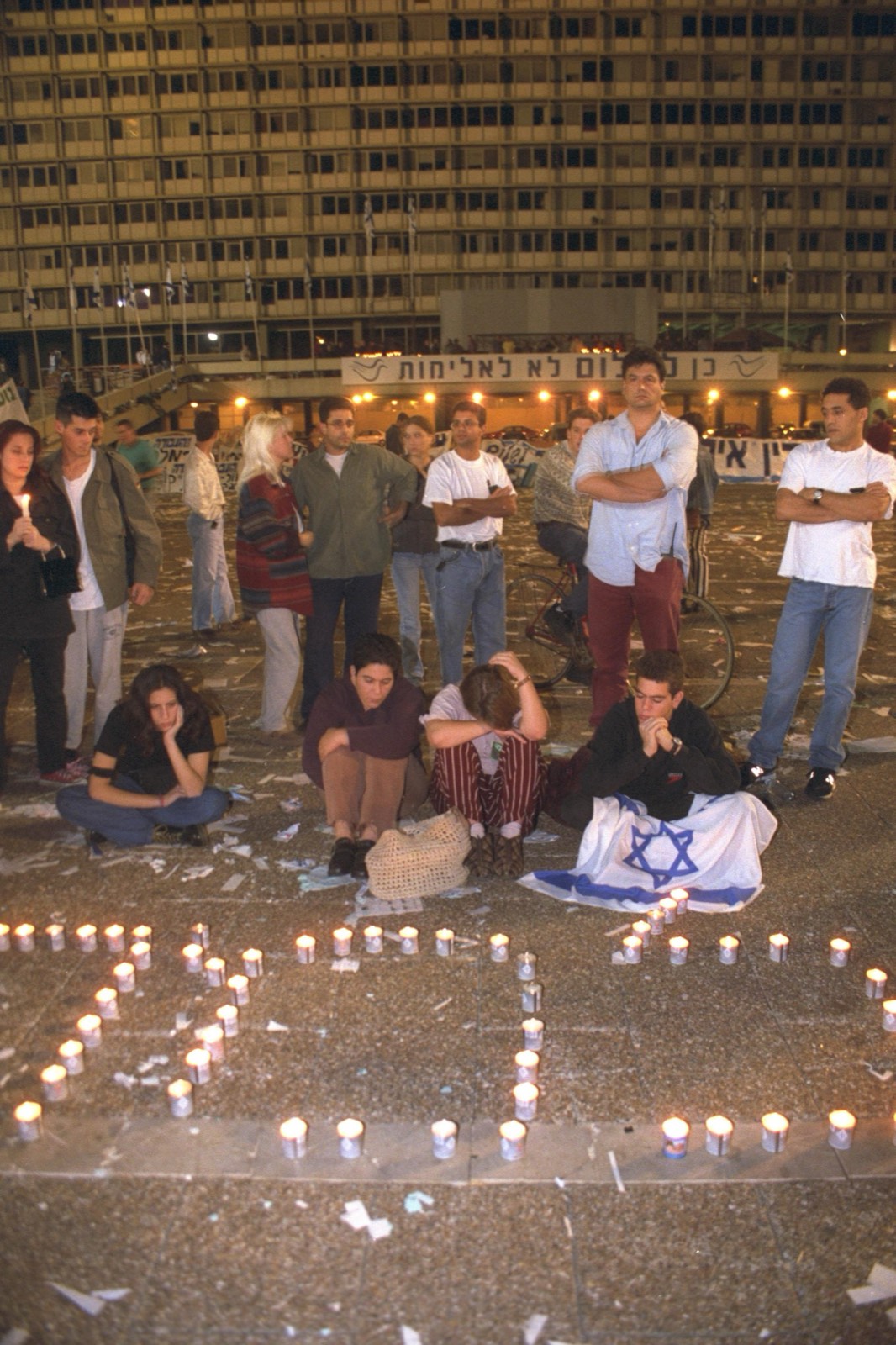 A night scene in Kings of Israel Square days after the assassination — young people standing and sitting on the paving among dozens of memorial candles arranged in Hebrew letters, one youth draped in an Israeli flag, the lit slab of Tel Aviv City Hall and a banner behind them.