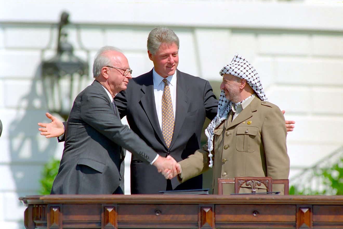 The signing ceremony of the Oslo Accords on the South Lawn of the White House, September 13, 1993 — U.S. President Bill Clinton stands at the centre with his arms spread, bringing together Israeli Prime Minister Yitzhak Rabin on the left and PLO Chairman Yasser Arafat on the right as they shake hands, with officials and flags behind them.