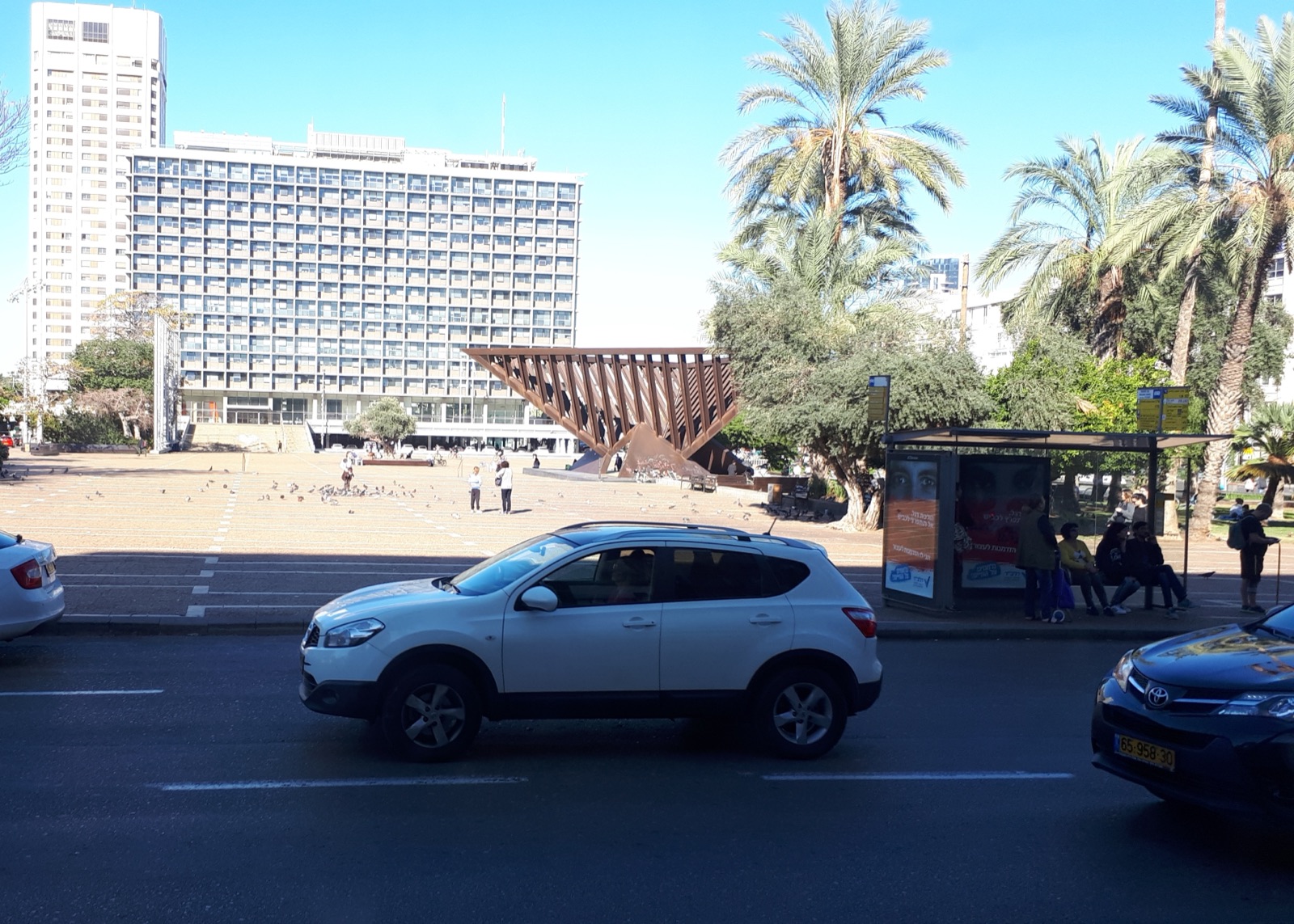 Kings of Israel Square — now Rabin Square — in Tel Aviv, photographed from across the street: a broad paved plaza with the long modernist slab of Tel Aviv City Hall behind it, the rust-coloured angular form of a memorial sculpture at its centre, and palm trees to the right.