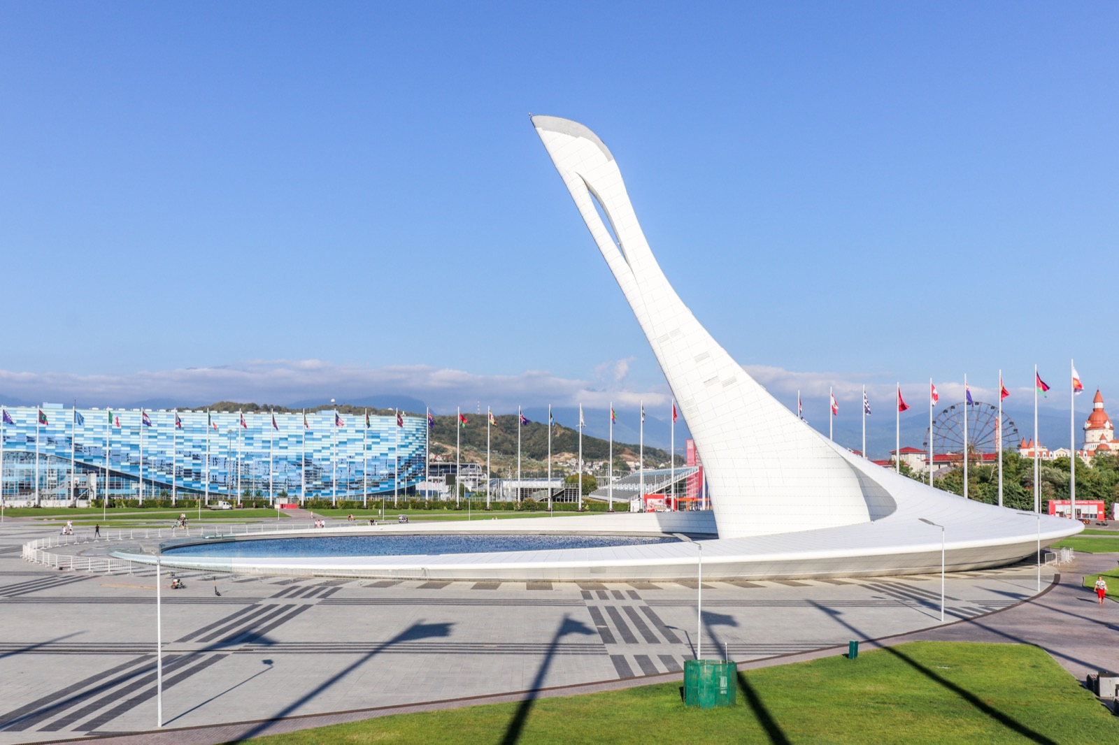 The Olympic cauldron at the Sochi 2014 Winter Olympics — a tall, curved white sculpture rising beside a circular reflecting pool in the Olympic Park, with rows of national flags and venues behind it under a clear blue sky.