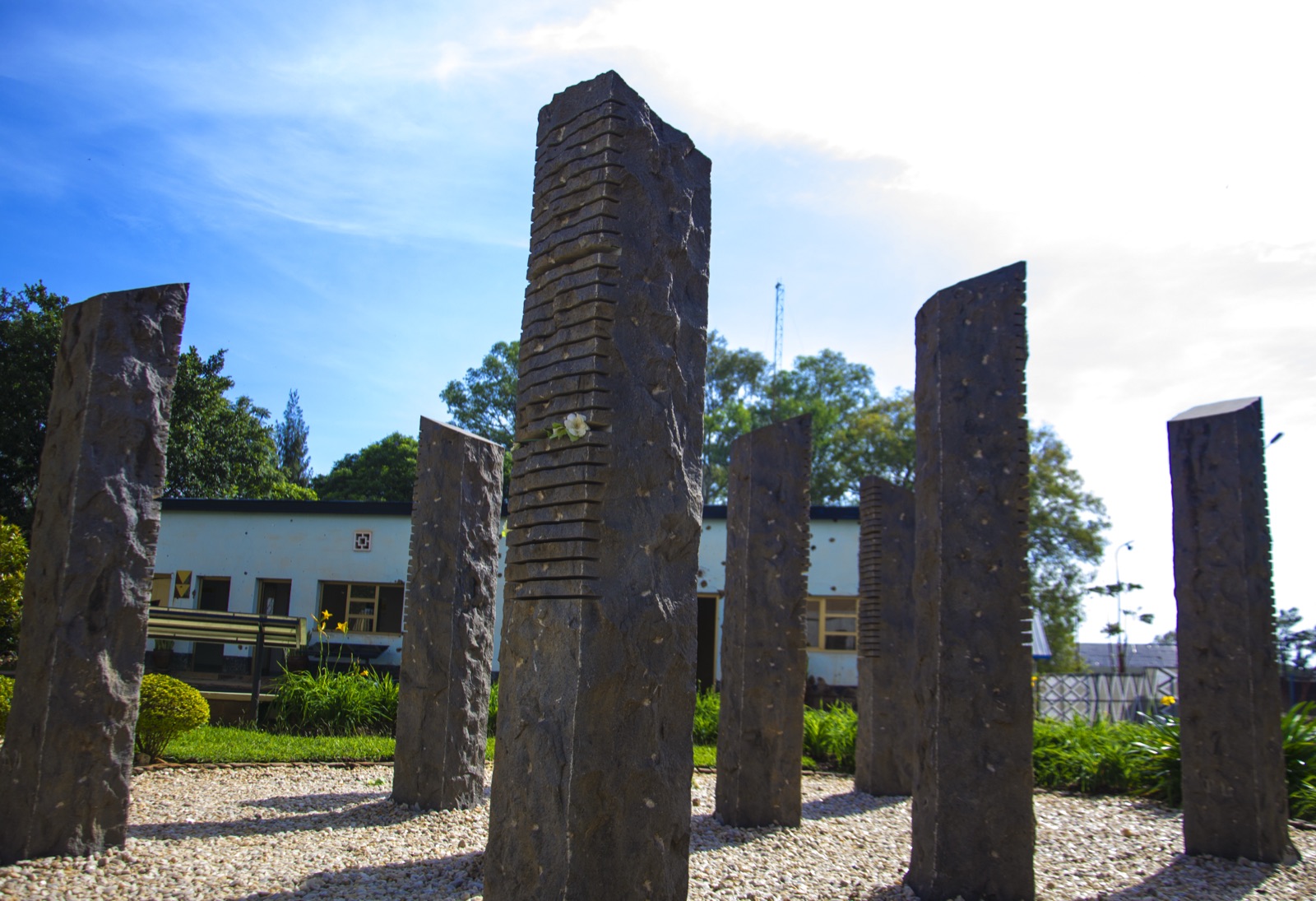 The Belgian Peacekeepers Memorial in Kigali — ten tall dark stone pillars of different heights standing in a gravel courtyard, each marked with horizontal cuts, with a low building and trees behind under a blue sky.