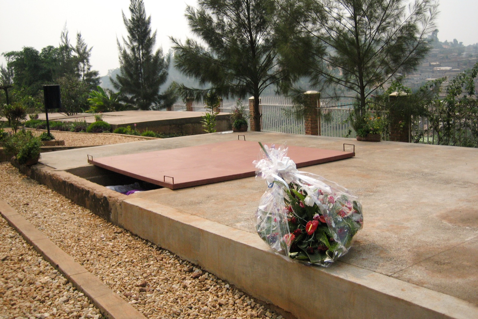 The grounds of the Kigali Genocide Memorial — a concrete mass-grave slab in a terraced garden with a bouquet of flowers laid beside it, trees and the hills of Kigali in the background.