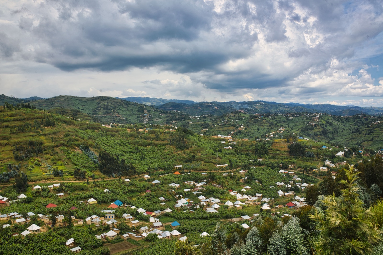 A wide view of the green, terraced hills of rural Rwanda — rolling farmland and scattered villages with metal roofs spread across steep slopes under a cloudy sky, the 'land of a thousand hills.'