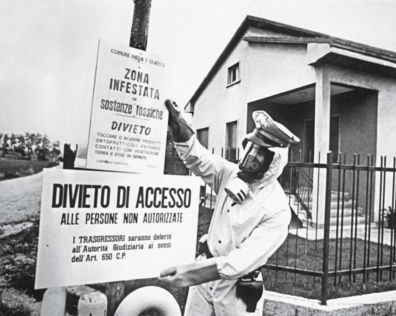 A carabiniere in a gas mask posting warning signs reading 'Zona infestata da sostanze tossiche' (zone contaminated by toxic substances) in front of a house near Seveso in 1976.