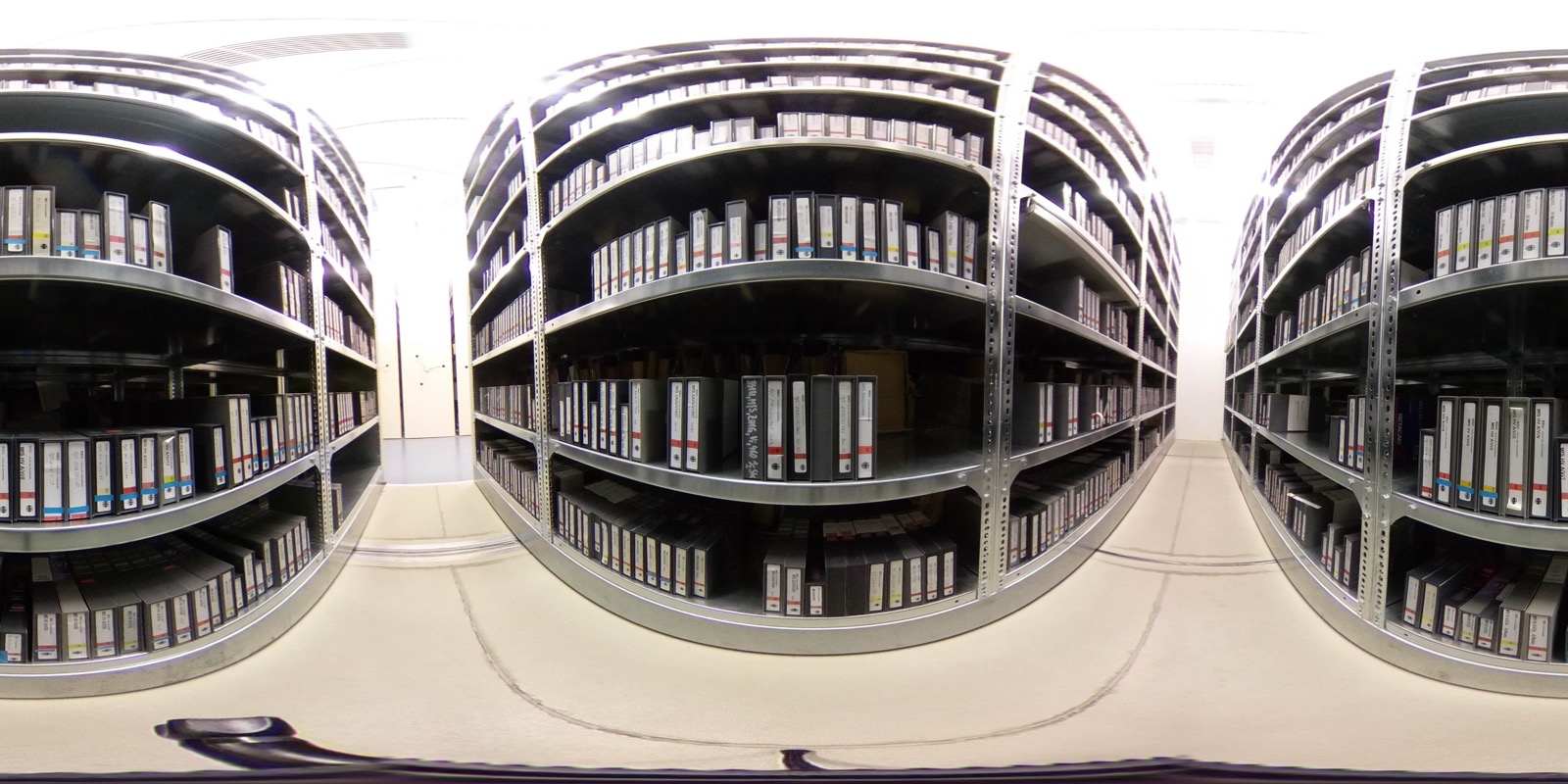 The Stasi Records archive shelving — long aisles of grey institutional shelving stacked floor-to-ceiling with cardboard files and binders, fluorescent lighting overhead, no people present.