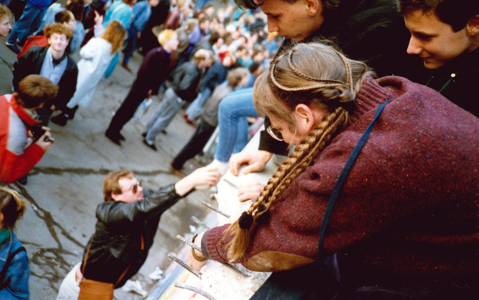 A West German crowd at the Berlin Wall on November 10, 1989 — people standing and seated on top of the wall near the Brandenburg Gate at night, some hammering at the wall, others embracing, brick-and-concrete wall in foreground, lit by floodlights and camera flashes.