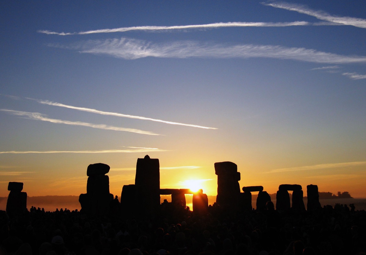 The sun rising over the stones of Stonehenge at the summer solstice, with crowds gathered.