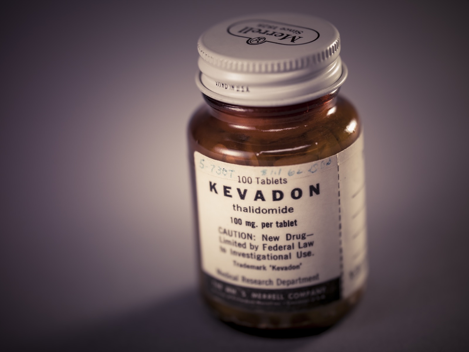 A small amber glass bottle from 1960 labelled 'KEVADON — thalidomide — 100 mg per tablet,' with the caution 'New Drug — Limited by Federal Law to Investigational Use,' from the Wm. S. Merrell Company, photographed against a dark background.