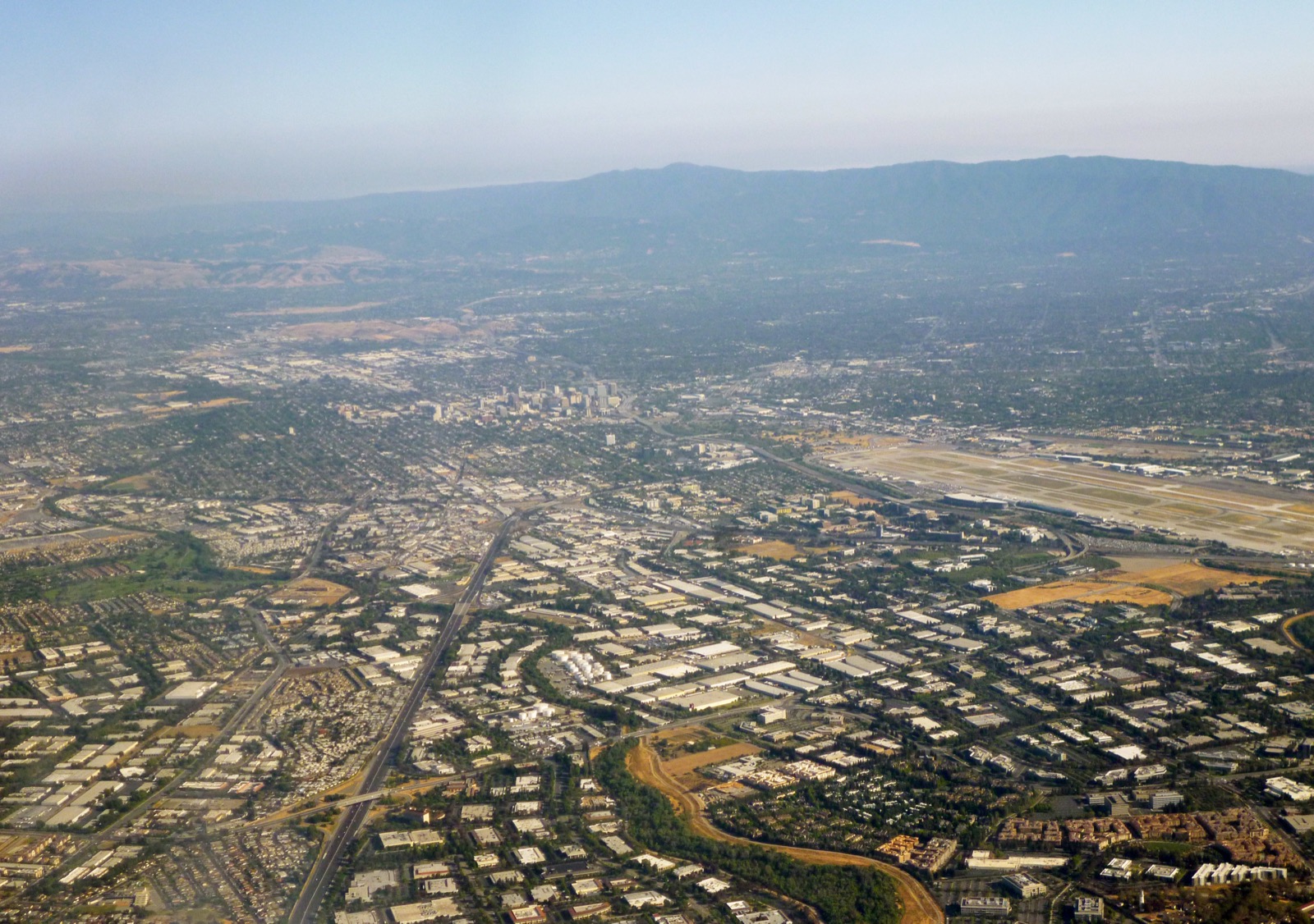 An aerial view of Silicon Valley looking south toward downtown San Jose, California — a sprawl of low buildings, roads, and greenery under a hazy sky, with hills in the distance.