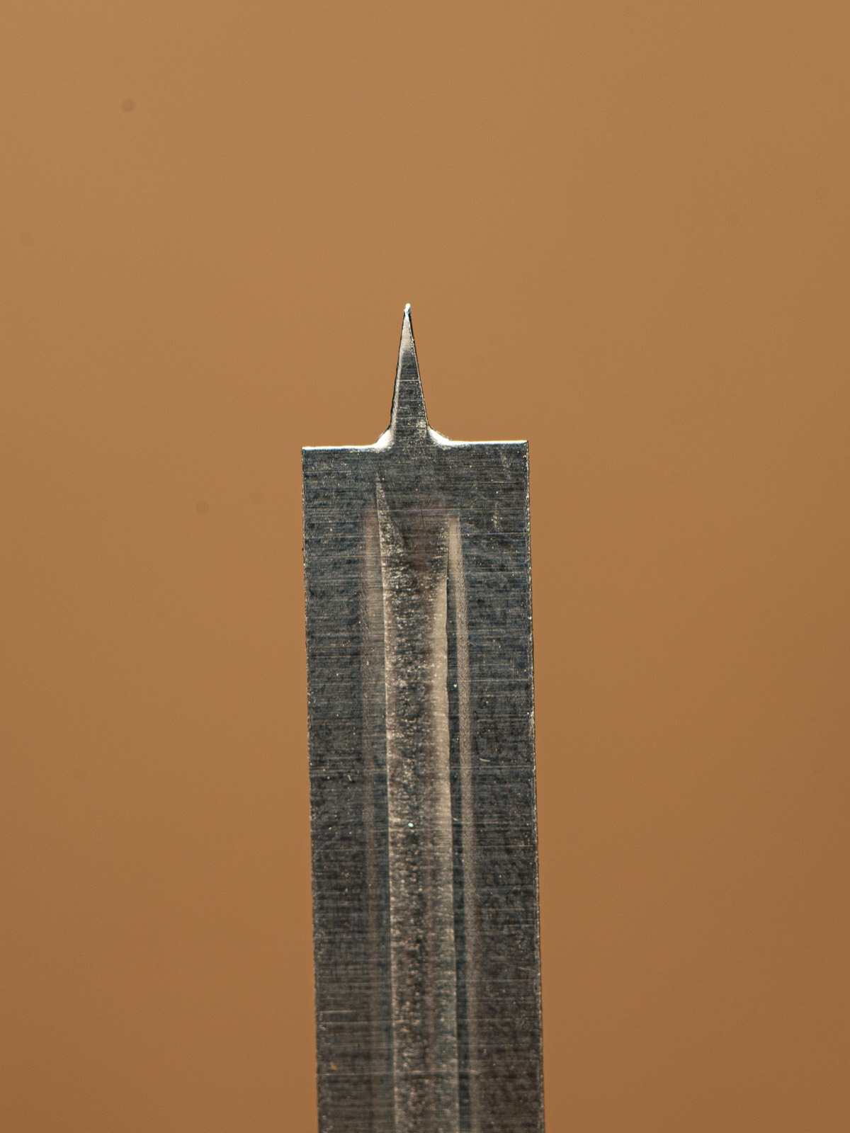 A close-up macro photograph of a lancet, the small sharp metal blade used to prick a fingertip to draw a drop of blood, against a plain background.