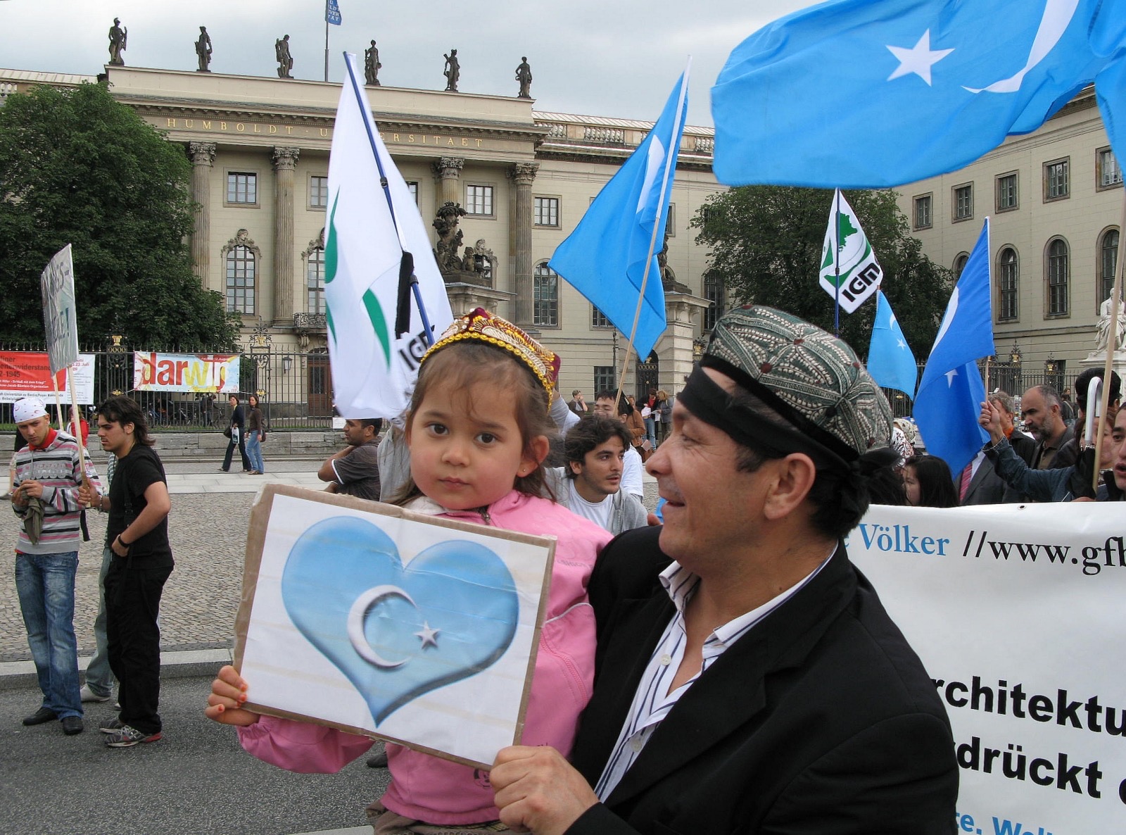 A Uyghur man wearing a traditional embroidered cap holds a young girl at a diaspora demonstration in Berlin; the child holds a hand-drawn sign with a light-blue heart bearing the white star-and-crescent of the East Turkestan flag, while pale-blue flags fly behind them in front of a grand university building.