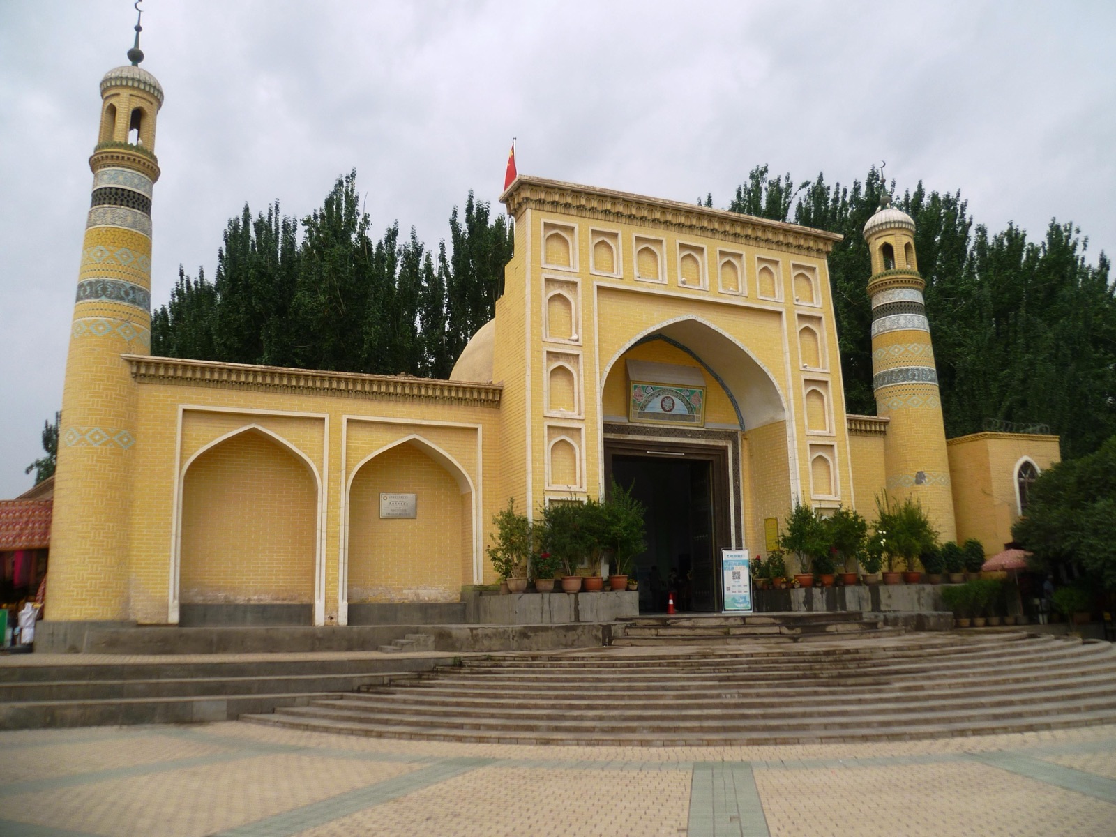 The Id Kah Mosque in Kashgar, a large mustard-yellow mosque with two minarets and an arched central portal, set behind a paved plaza under a blue sky.