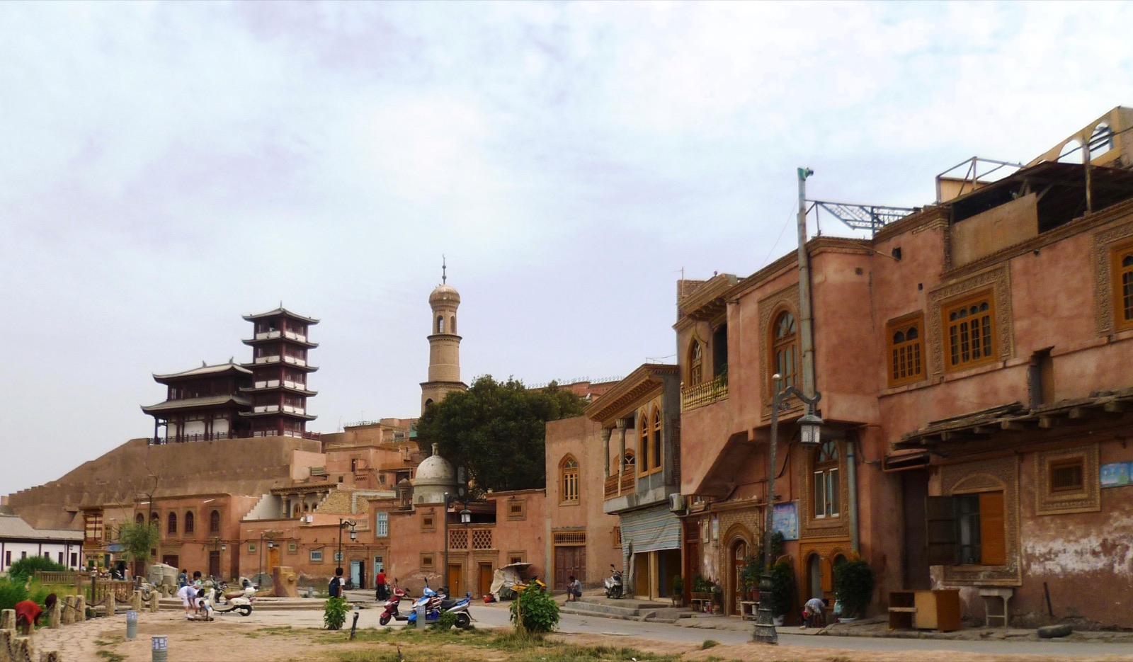 A street in the old city of Kashgar lined with traditional mud-brick buildings and shopfronts, a few people walking under a clear sky.