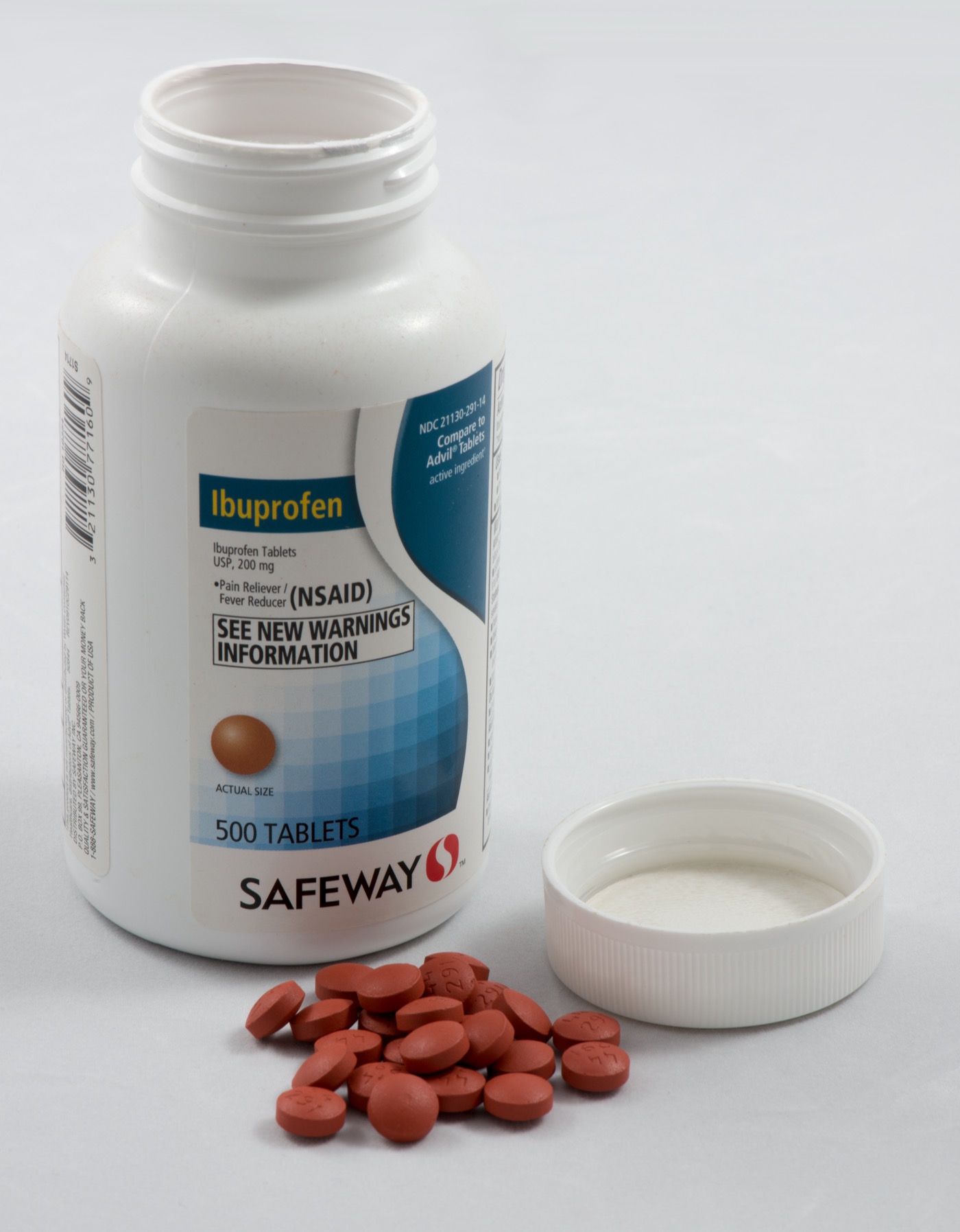 A bottle of ibuprofen tablets with the cap removed, several round white tablets spilled in front of it on a plain surface.