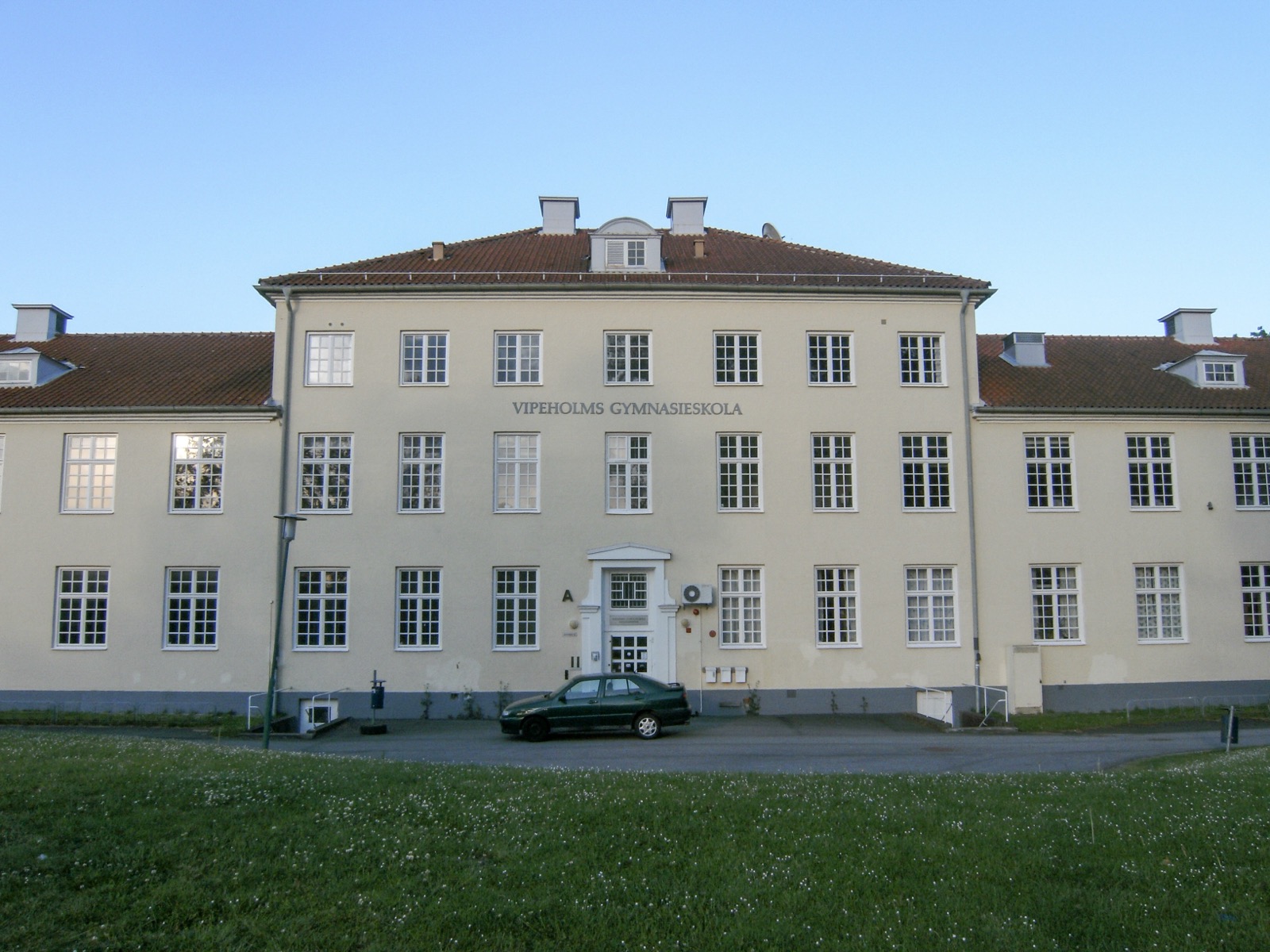 The main building of the former Vipeholm hospital in Lund, Sweden — a long, pale-yellow three-storey institutional building with a red-tiled roof and rows of windows, now bearing the words 'Vipeholms Gymnasieskola' above the entrance, seen across a lawn.