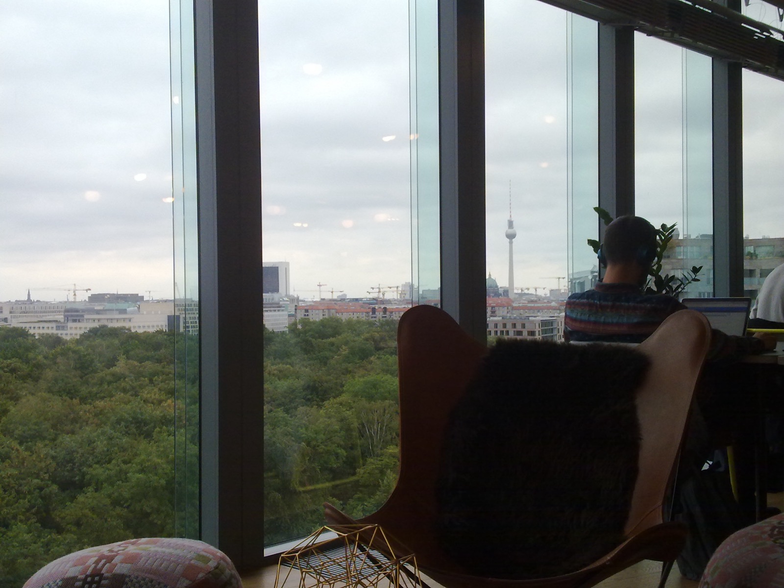 The interior of a WeWork coworking space in Berlin, with floor-to-ceiling windows overlooking the city and its TV tower, designer butterfly chairs, plants, and a person working on a laptop with headphones on.