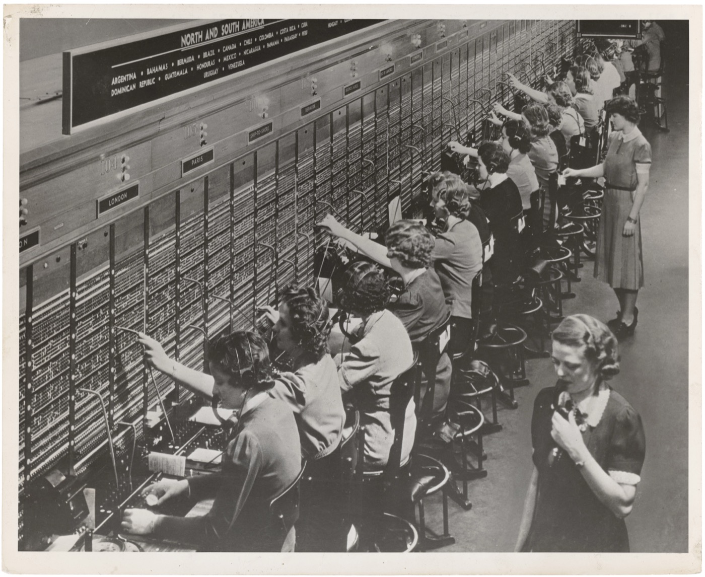 A historic photograph of women working at a Bell System telephone switchboard, seated in a long row before the equipment.