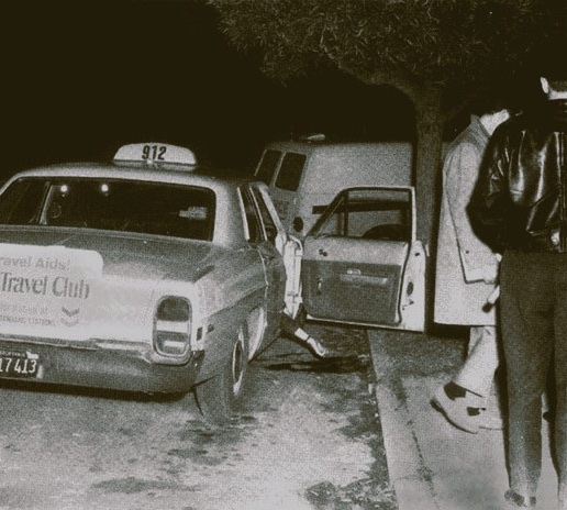 A black-and-white night-time police crime-scene photograph of a 1960s Yellow Cab, number 912, parked at the kerb in Presidio Heights with its rear door open, investigators in suits standing beside it under a tree.